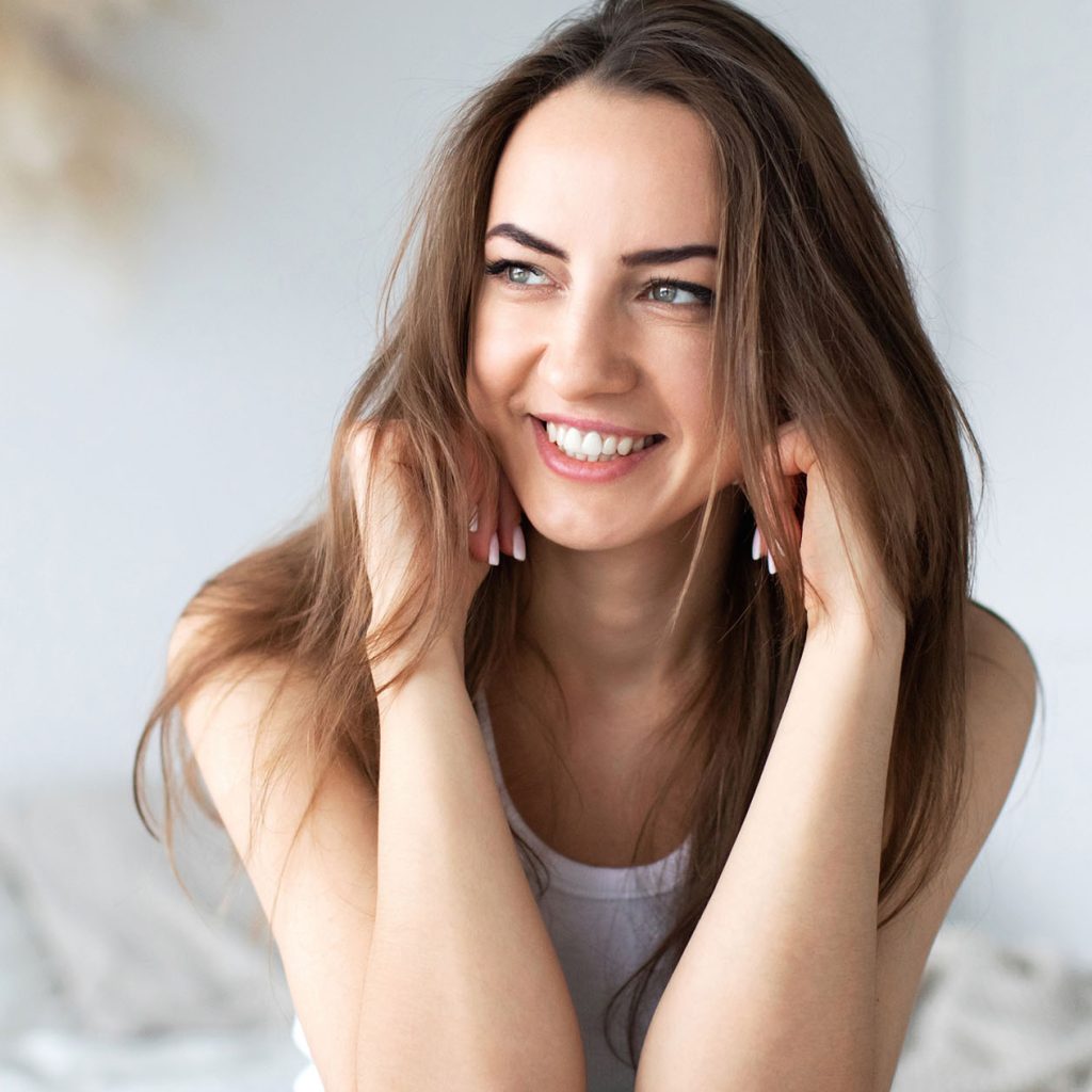 Young lady with brunette hair smiling with exposed teeth against a light coloured background